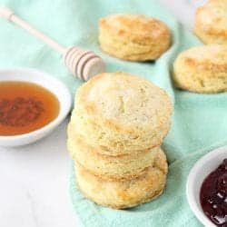 Stack of biscuits on green napkin with honey and jam beside them.
