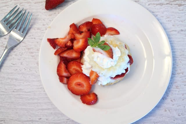 Overhead of dessert on a white plate surrounded by chopped strawberries with 2 forks beside it.