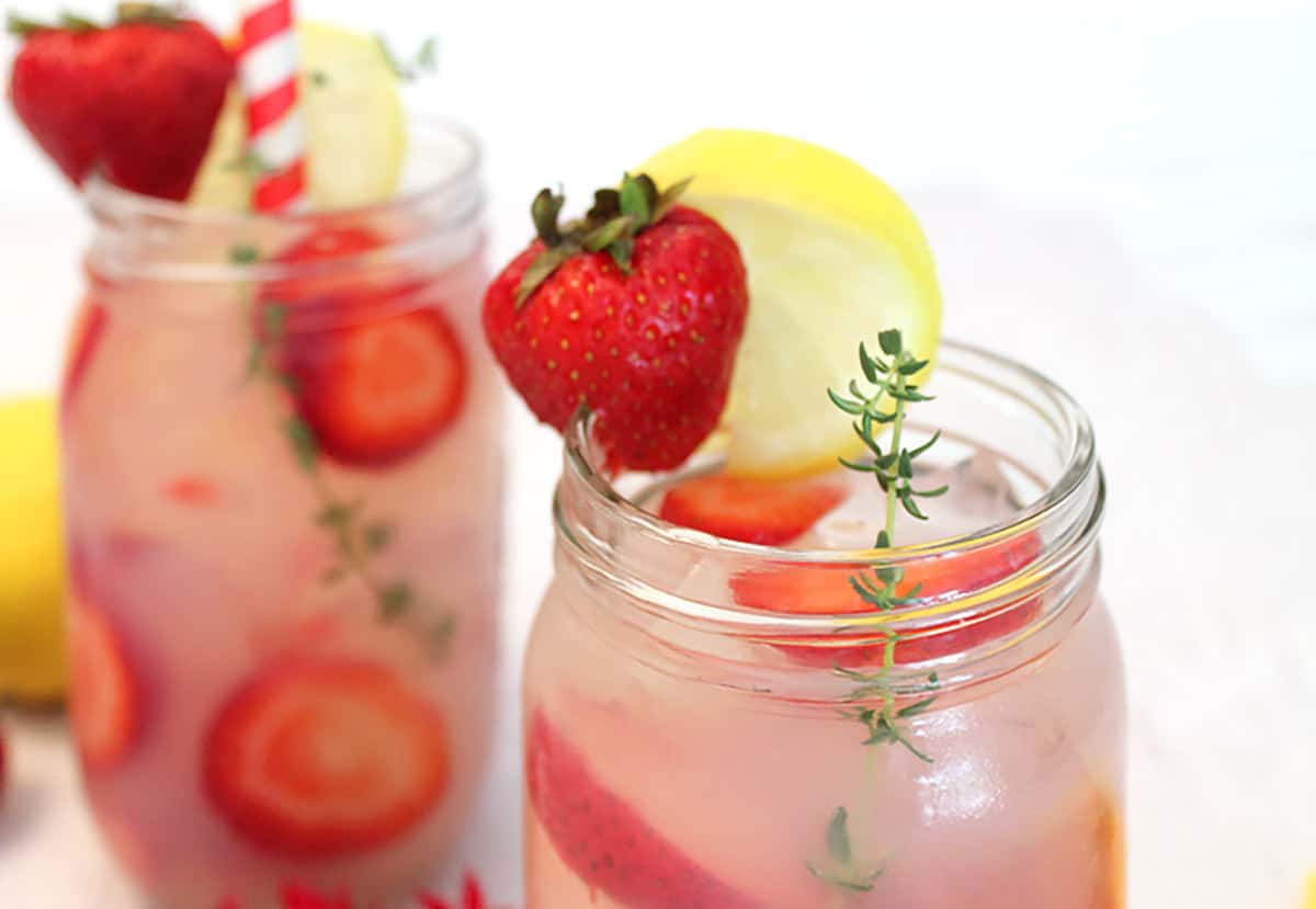 Closeup across lip of mason jar showing pink colored lemonade with strawberry, lemon slice and thyme garnish.