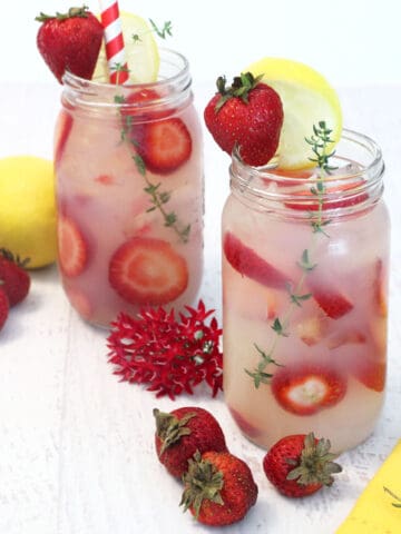 2 glasses of vodka lemonade showing sliced strawberries floating in drink with strawberries and lemon beside them.