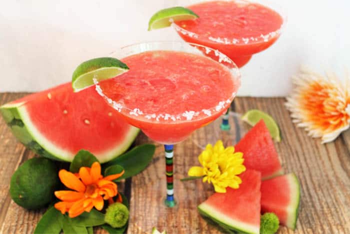 2 margarita glasses filled with frozen watermelon margarita with slice of lime and salt rim amid watermelon slices and flowers.