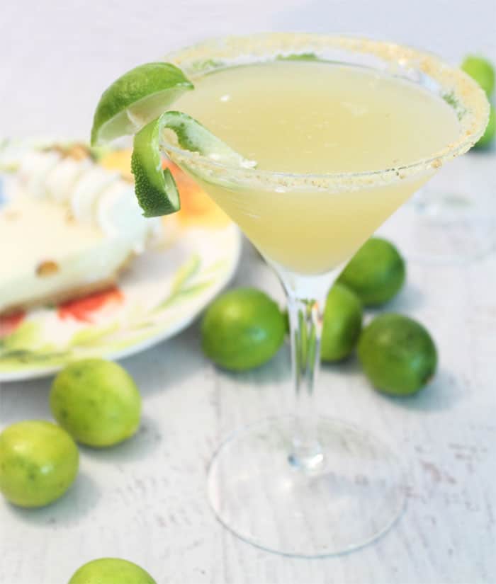 A single Key Lime Pie Martini with lime garnish on white table with key lime pie behind it.