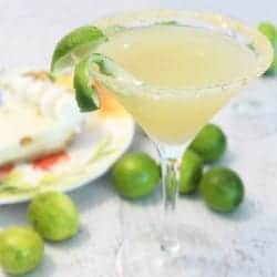 Martini glass with graham cracker rim, filled with key lime martini and surrounded by key limes.