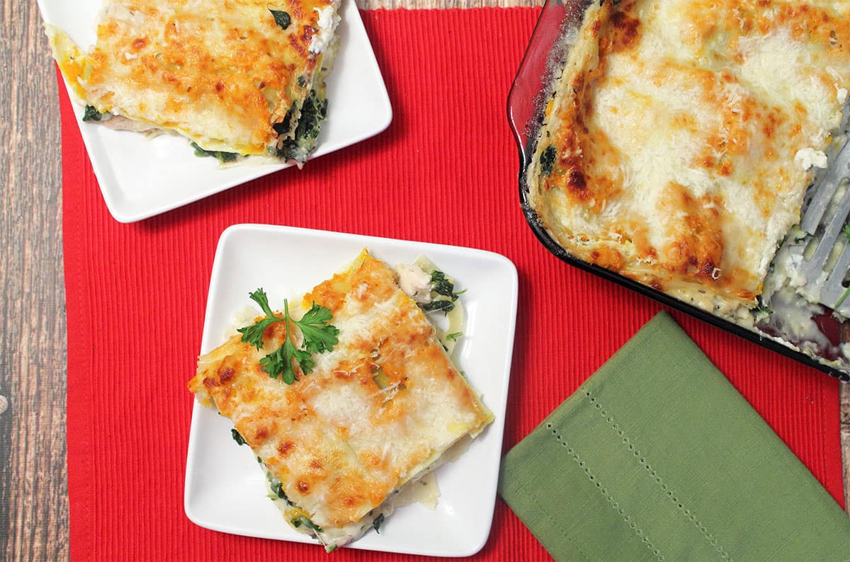 Overhead of 2 servings of lasagna on white plates on red placemat with casserole on side.