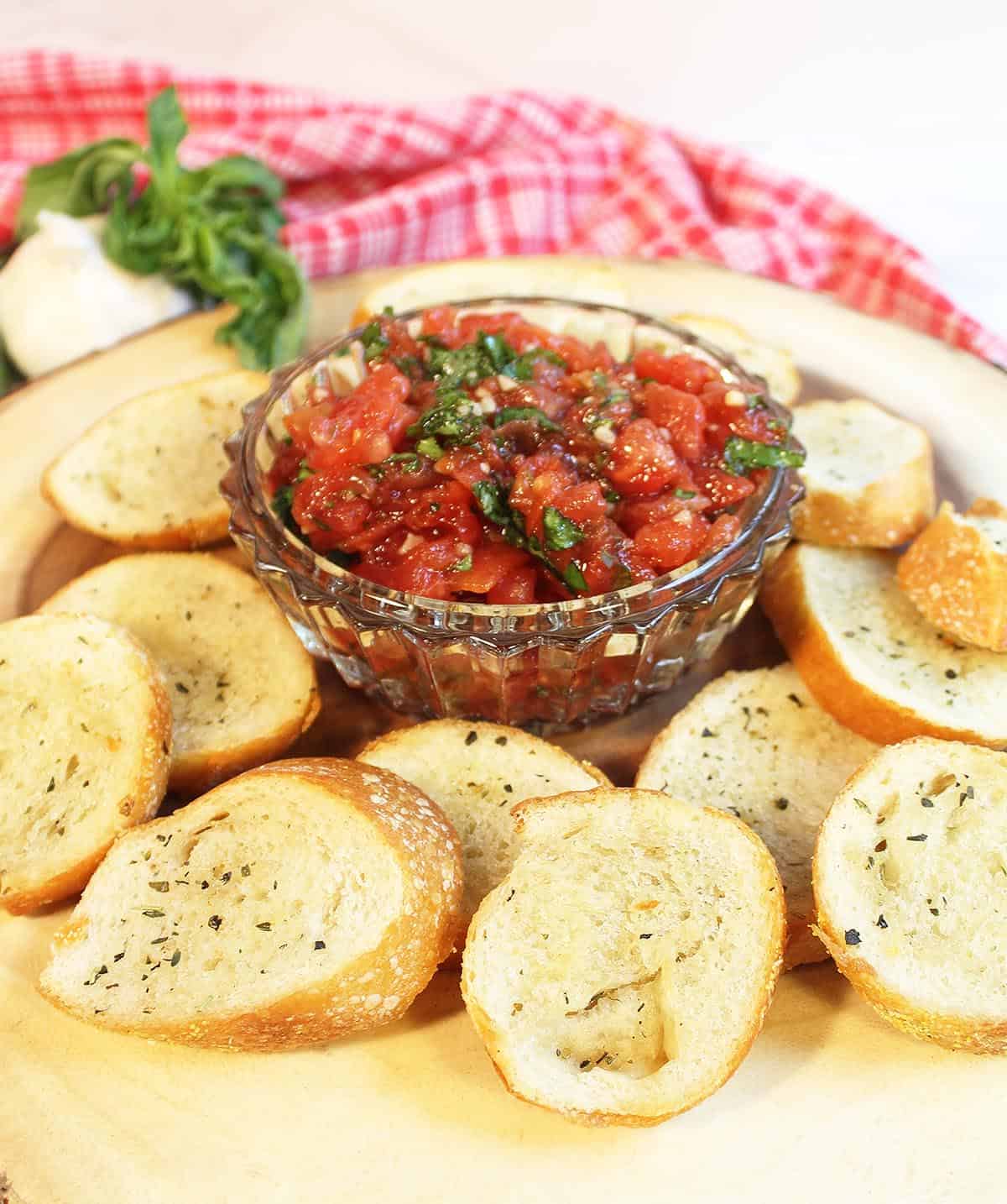 Bruschetta in crystal bowl on wood circle with bread crisps around it.