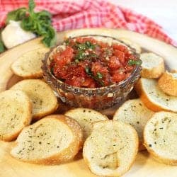 Bruschetta in crystal bowl on wood circle with bread crisps around it.