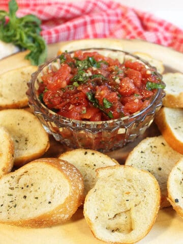 Bruschetta in crystal bowl on wood circle with bread crisps around it.