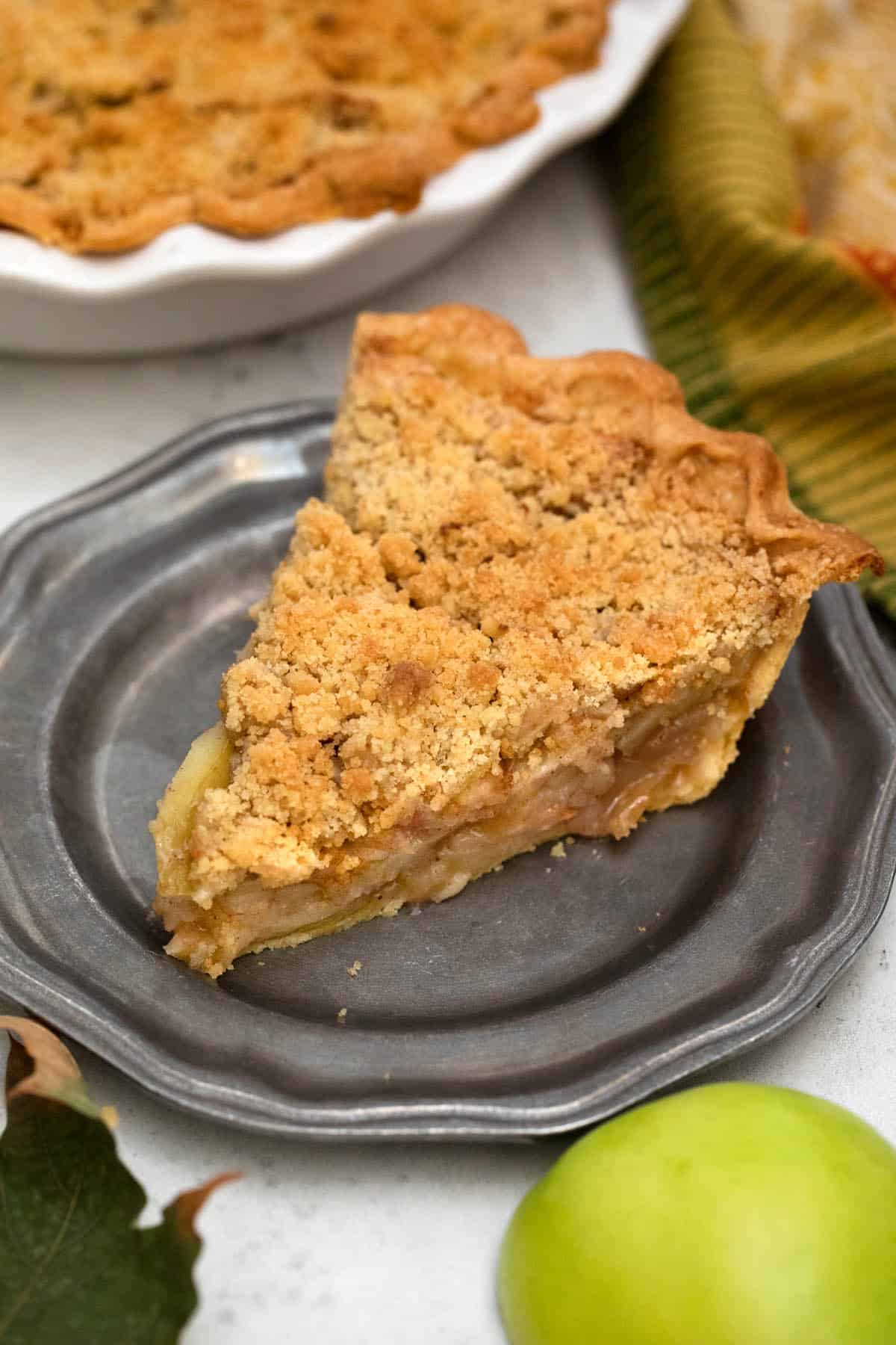 Slice of pie on pewter plate with apple in front.