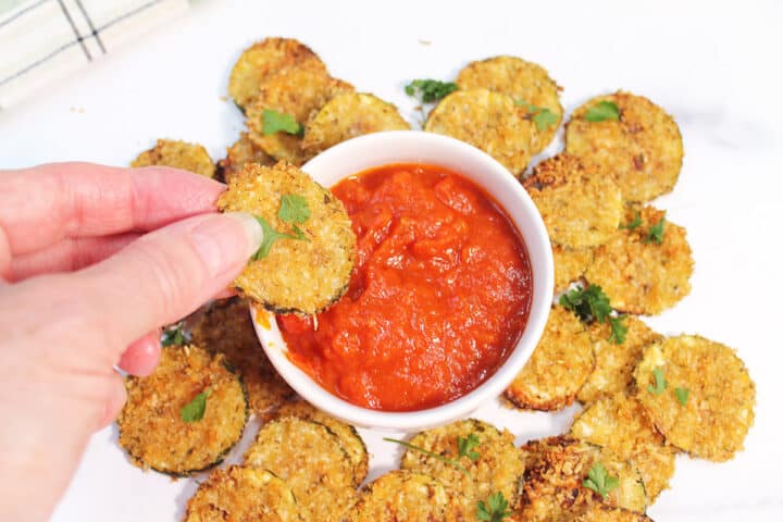 Dipping zucchini chip in marinara with chips surrounding bowl.