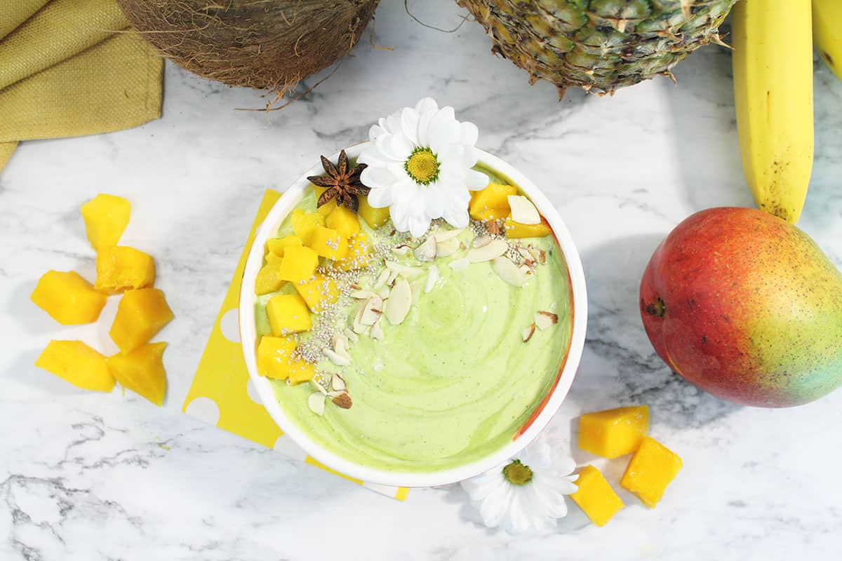 Spinach pineapple smoothie in bowl on white table with mango pieces around bowl.