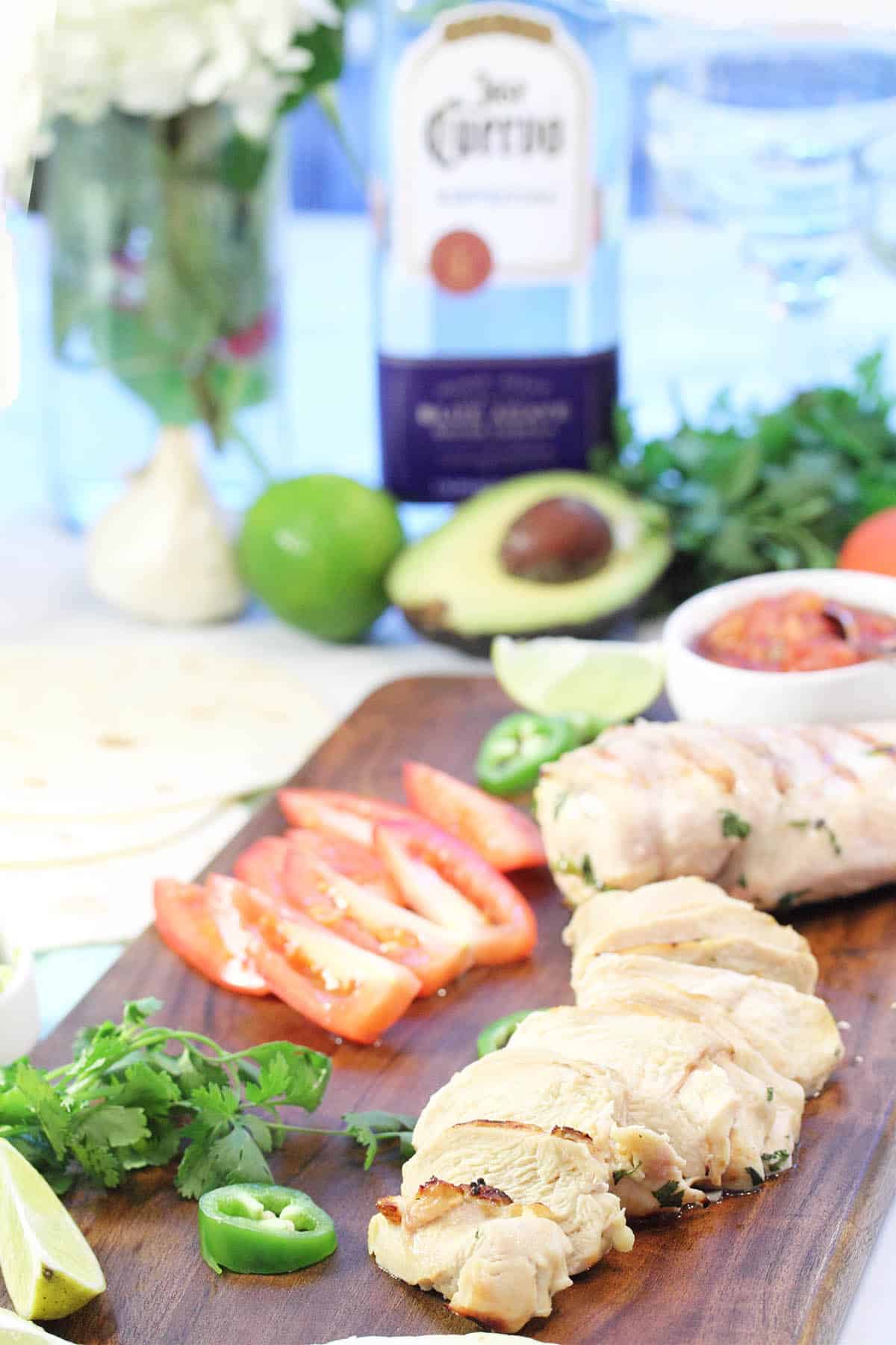 Chicken dinner on wooden board with avocado, lime and tequila behind it.