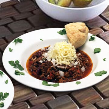 Bowl of hearty chili topped with cheese on brick surface.