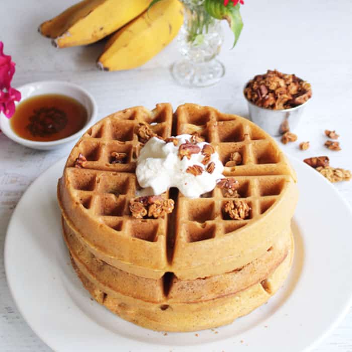 Stacked waffles on a white plate with whipped cream on top and syrup and bananas in background.