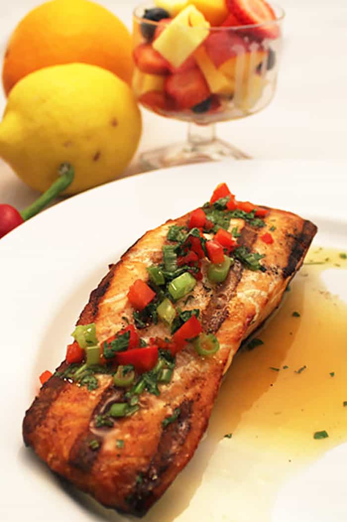 Grilled Salmon drizzled with orange sauce on white plate with fruit in background.