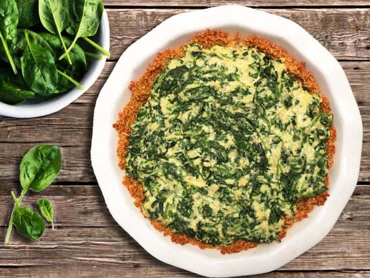 Spinach pie in white pie dish on wooden table with spinach in bowl beside it.