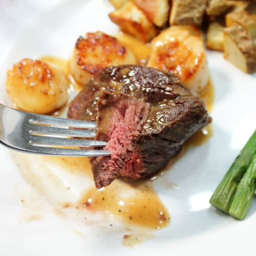 Holding bite of steak over plate of pan seared steak and scallops.