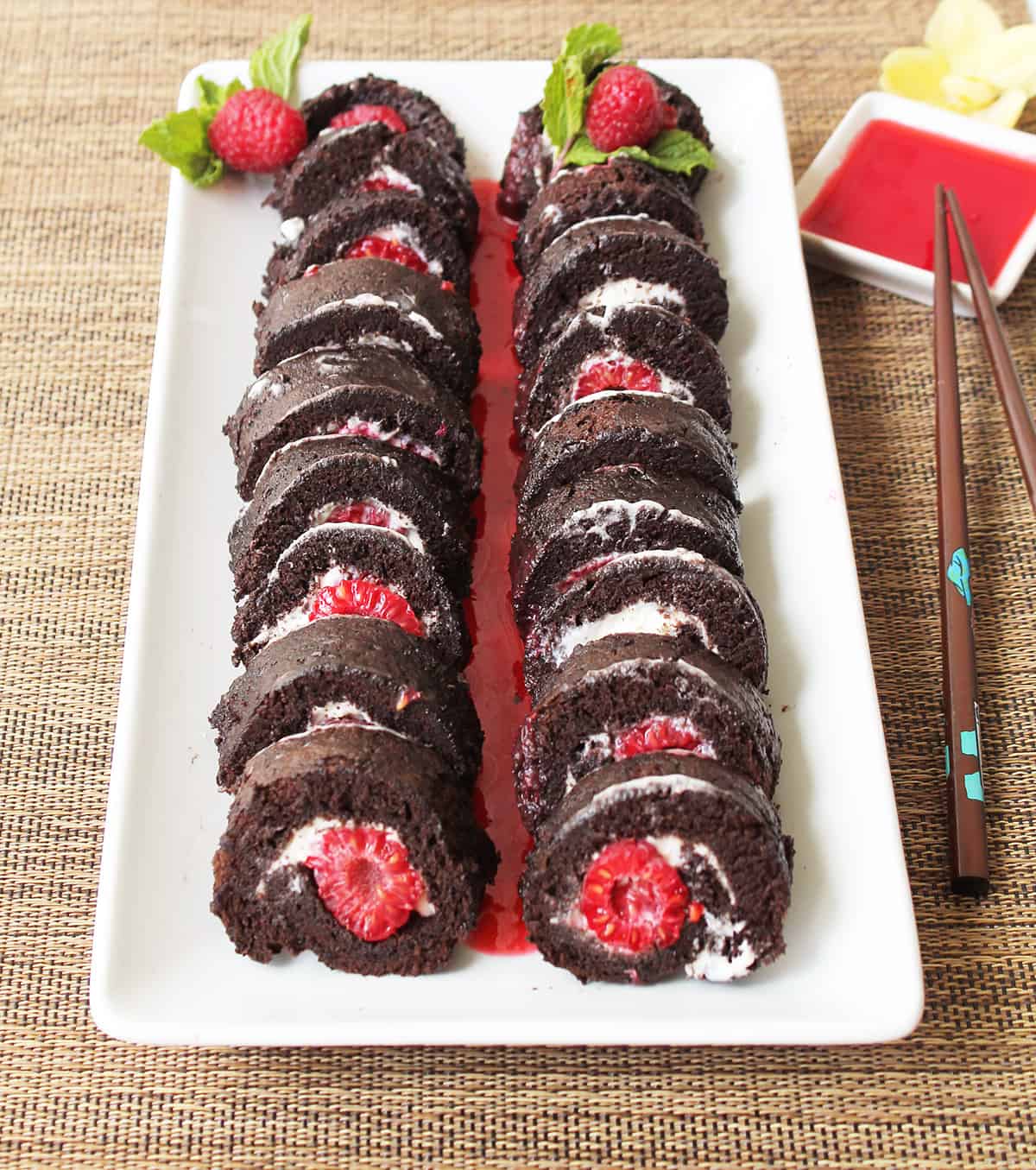 Two chocolate raspberry sushi rolls on white platter with raspberry sauce.