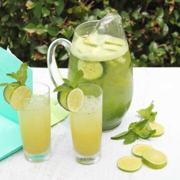 Pitcher of Cucumber Cocktails with two filled glasses in front and lime and mint on table.