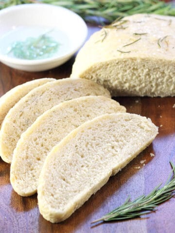 Partially sliced rosemary bread on wooden serving board with dipping bowl and rosemary.