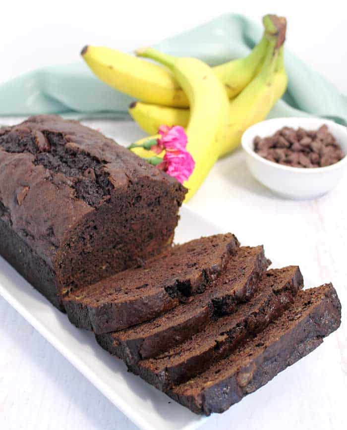 Sliced banana bread on white plate with bananas and chocolate chips in background.