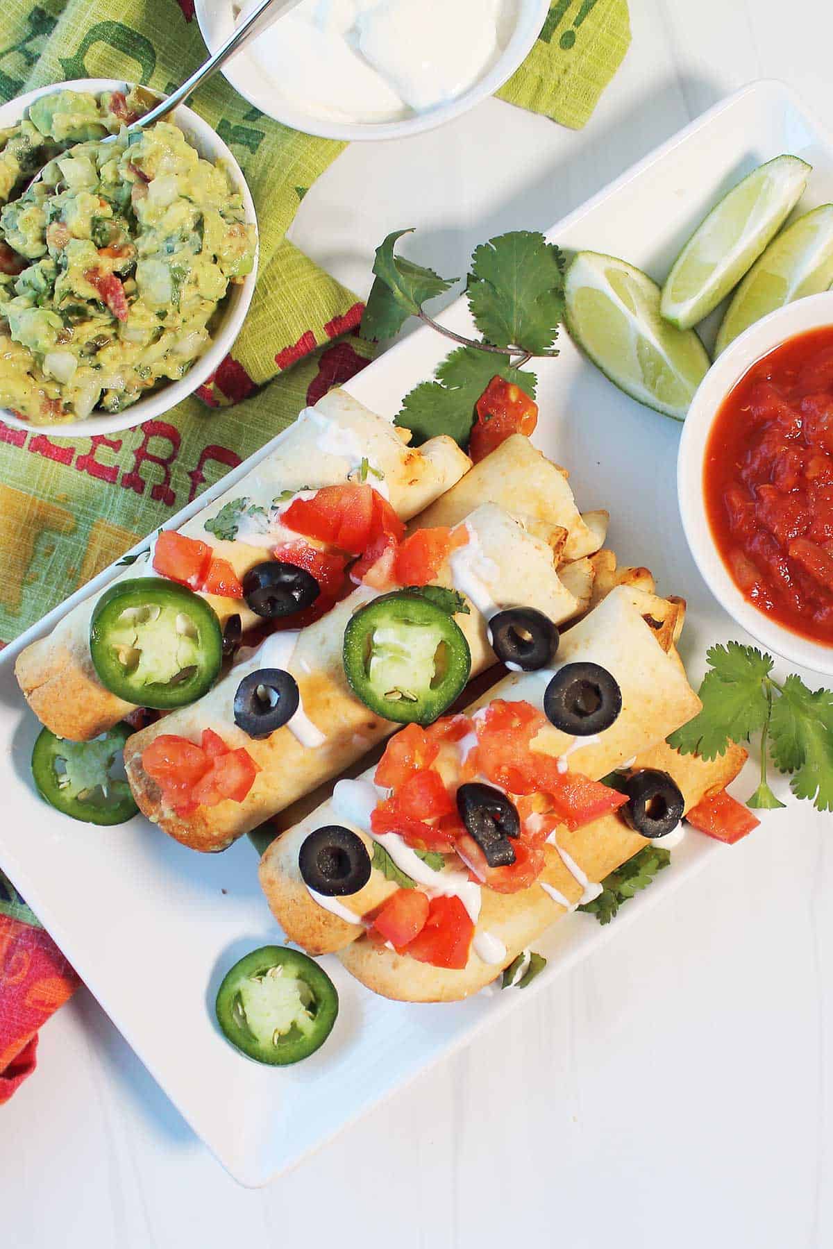 Overhead of garnished air fryer chicken taquitos with sides of guacamole, salsa and sour cream.