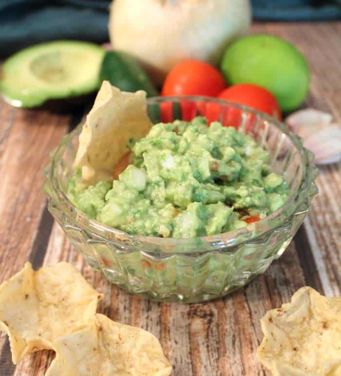 Guacamole in crystal bowl with ingredients in background and chips in front.