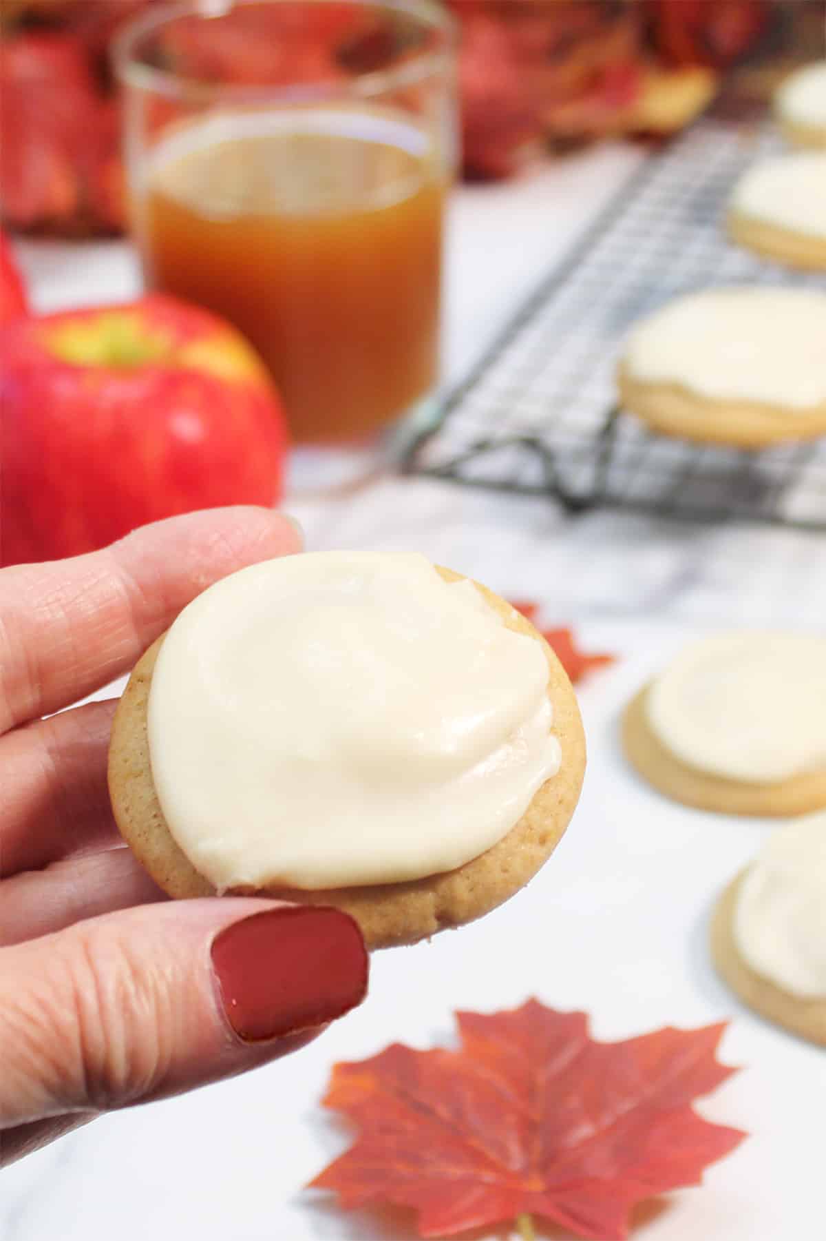 Holding a frosted apple cookie over white table with cider in background.