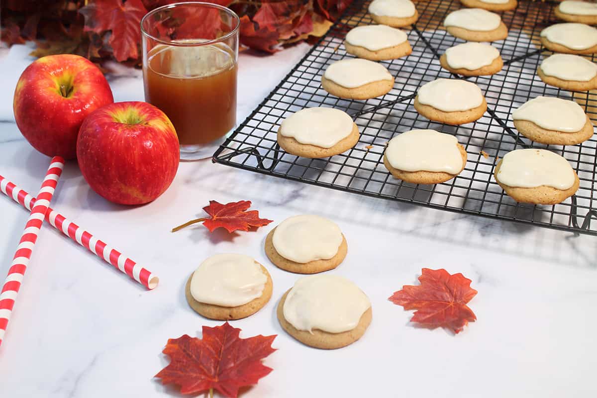 Frosted cookies on wire rack with three in front and apples and fall leaves by them.