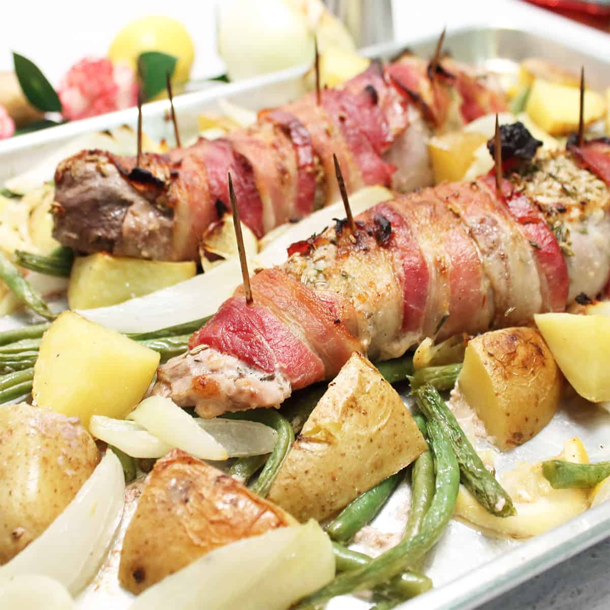 Closeup of pork tenderloin wrapped in bacon with vegetables on sheet pan.