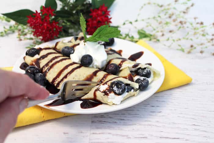 Cutting into the blueberry crepes set on a white plate with yellow napkin.