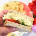 Holding half egg salad sandwich over plate.