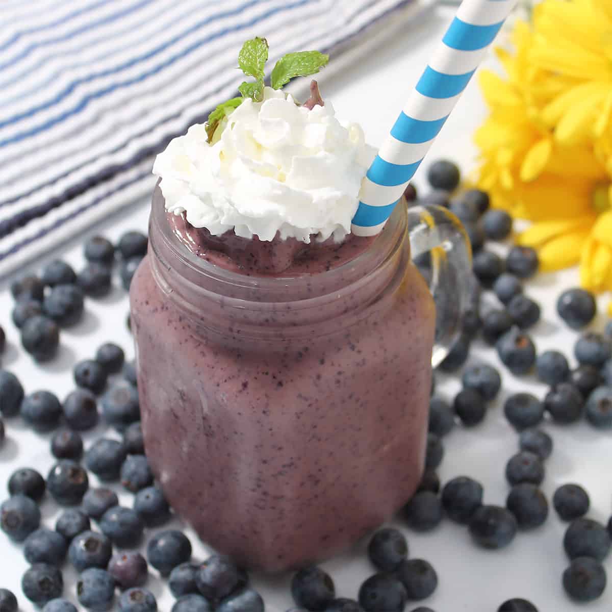 Blueberry Coffee Smoothie garnished with whipped cream amid blueberries with yellow flowers in background.