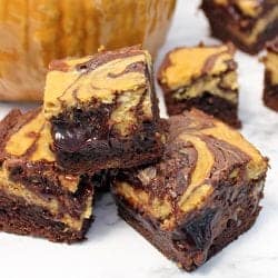 Stacked pumpkin cheesecake brownies on white table in front of pumpkin.