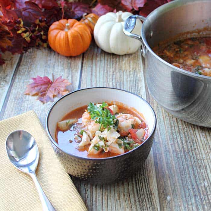 Serving of fish stew in brown bowl on wooden table with pot of stew in background.