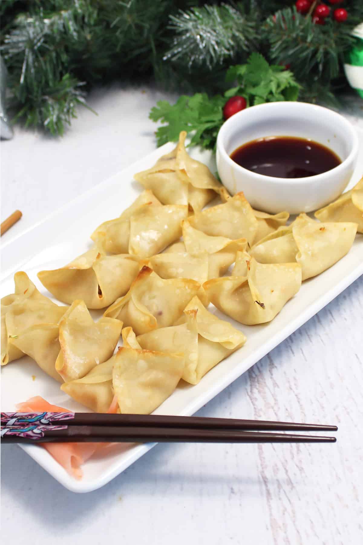 Baked chicken wontons on white platter with dip and greenery in back.