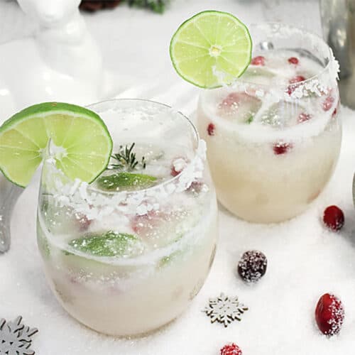 Closeup of two white christmas margaritas with lime wedge and sugared cranberries around them.