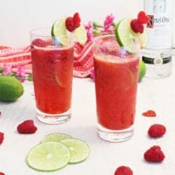 Two raspberry lime rickey cocktails with garnish on white table with raspberries and lime slices by them.