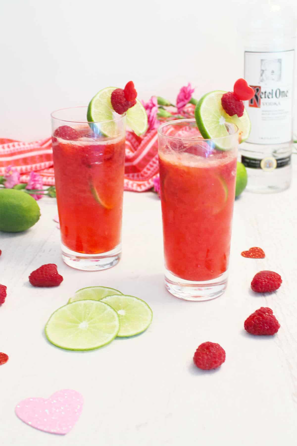 Two raspberry lime rickey cocktails with garnish on white table with raspberries, lime slices and vodka bottle.