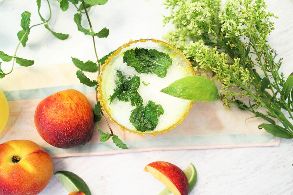 Overhead of single cocktail with gold sugar rim lime garnish and peach lime wedges and mint leaves around it.