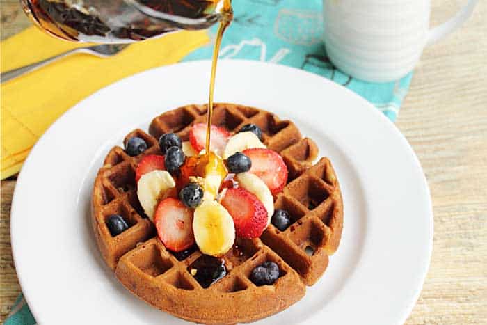 Pouring maple syrup over Banana Bread Waffles on white plate.