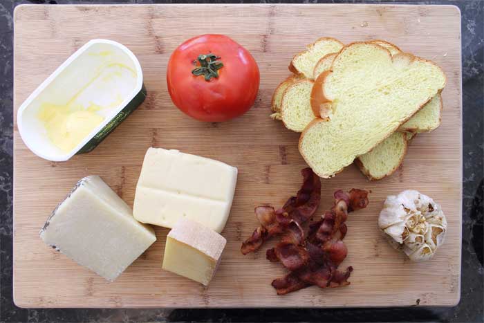 Ingredients for sandwich on wooden cutting board including roasted garlic and bacon.