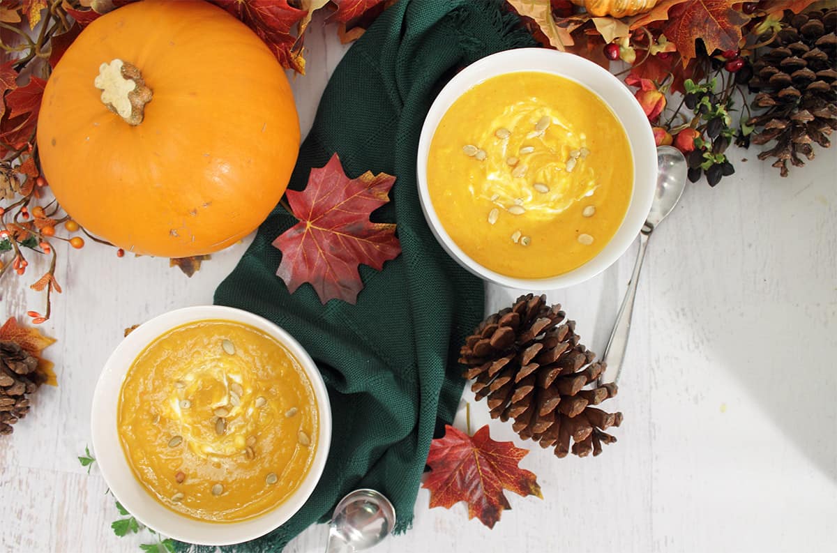 Overhead of two bowls of squash soup on dark green napkin  next to pumpkin.