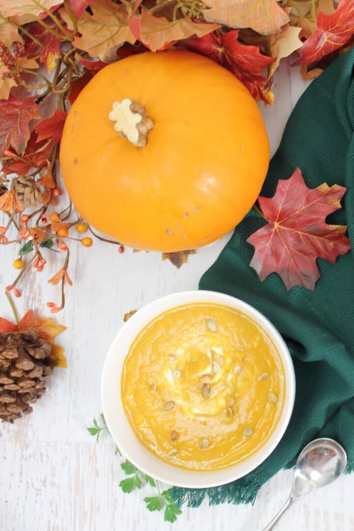 Overhead of soup in white bowl next to pumpkin, fall leaves and dark green napkin.