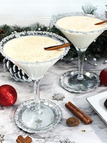 Two martini glasses filled with eggnog martini adorned with cinnamon sticks on white tablecloth.