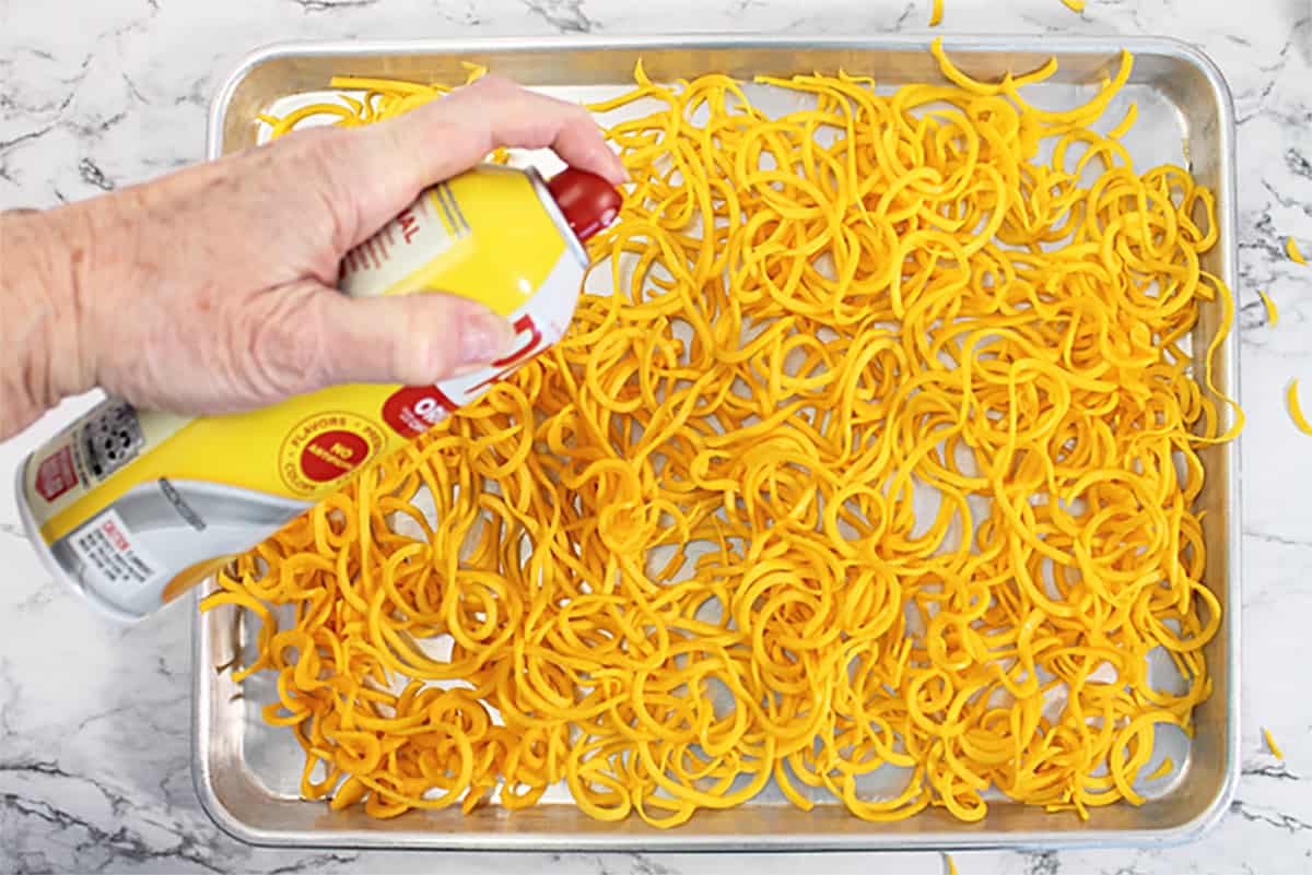 Spraying butternut squash noodles with vegetable spray on cookie sheet.