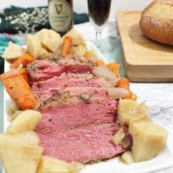 Siced corned beef surrounded by potatoes and vegetables on white platter with beer in background.