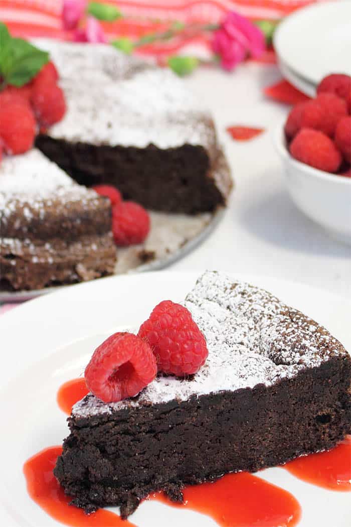 Closeup of chocolate torte on white plate drizzle with raspberry sauce. Full torte in background.