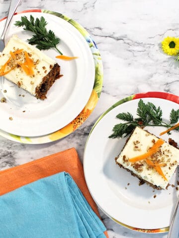 Overhead of two slices of carrot cake on white plates with orange and turquoise napkins.