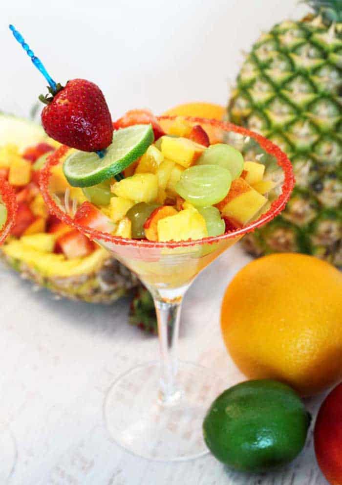 Fruit cocktail in martini glass with red sugar rim with fruit around it.