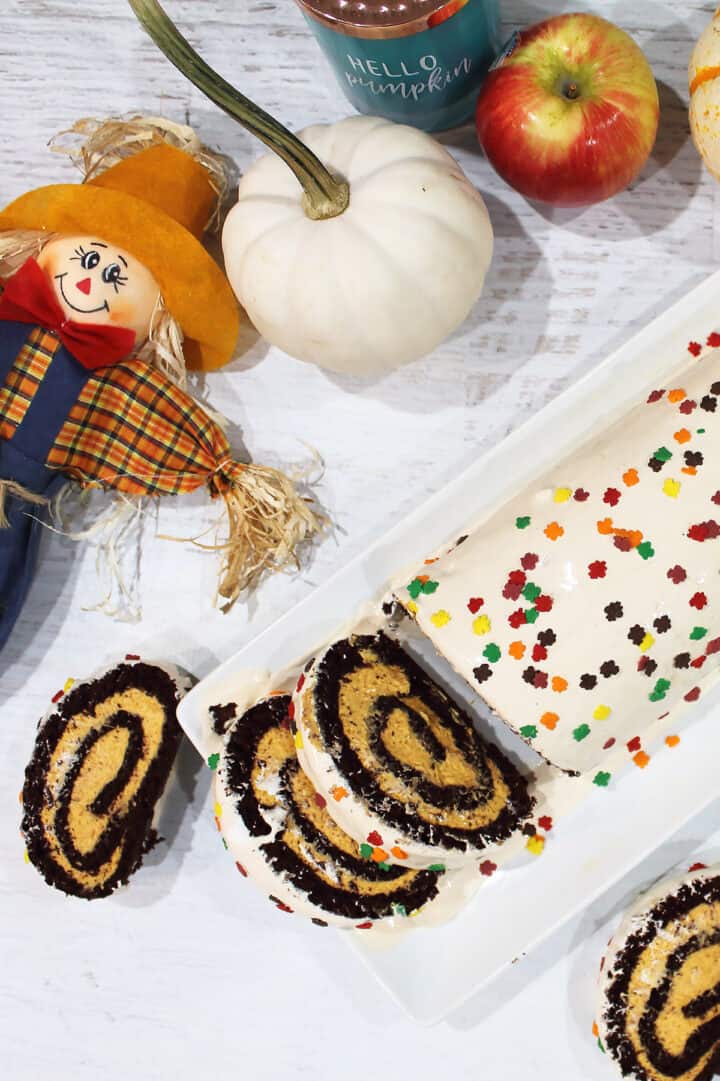 Sliced pumpkin cake roll with 2 slices on white table and fall pumpkin, apple and scarecrow around it.